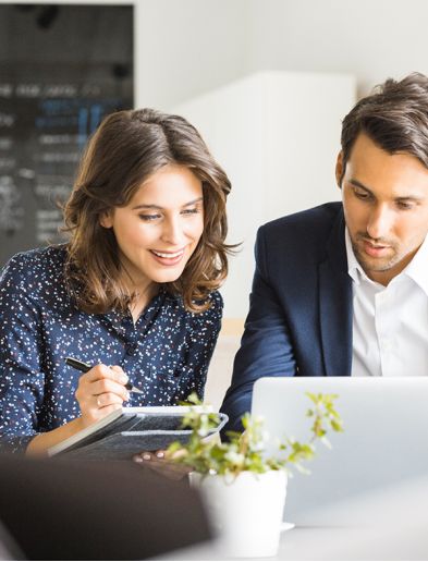 Job bei K-Plus Personalmanagement: Personalkauffrau mit Notizbuch und Personalkaufmann sitzen gemeinsam vor Laptop