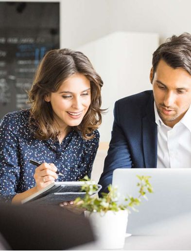 Job bei K-Plus Personalmanagement: Personalkauffrau mit Notizbuch und Personalkaufmann sitzen gemeinsam vor Laptop