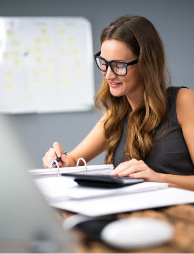 Job bei K-Plus Personalmanagement: Bürokauffrau mit Taschenrechner prüft Zahlen in Ordner